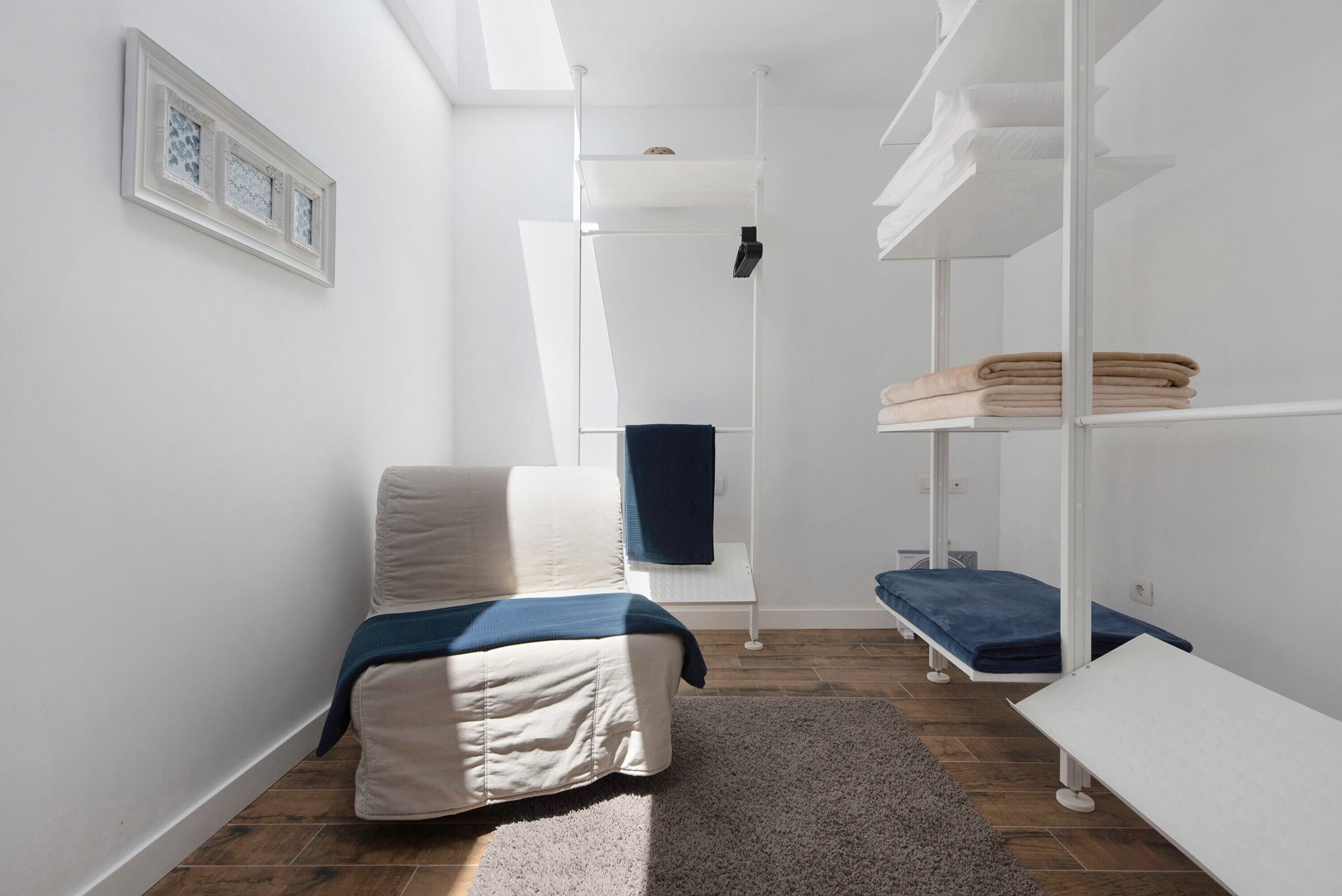A narrow room with a bed, a bench, and a shelf unit with folded towels.