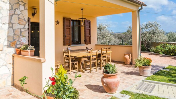 Un patio exterior techado con mesa y sillas de madera, paredes de piedra y plantas en macetas.