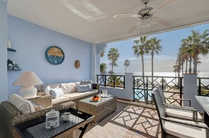 A room with a sofa, coffee table, and wicker furniture, a clock on the wall, and a balcony with a view of palm trees and the ocean.