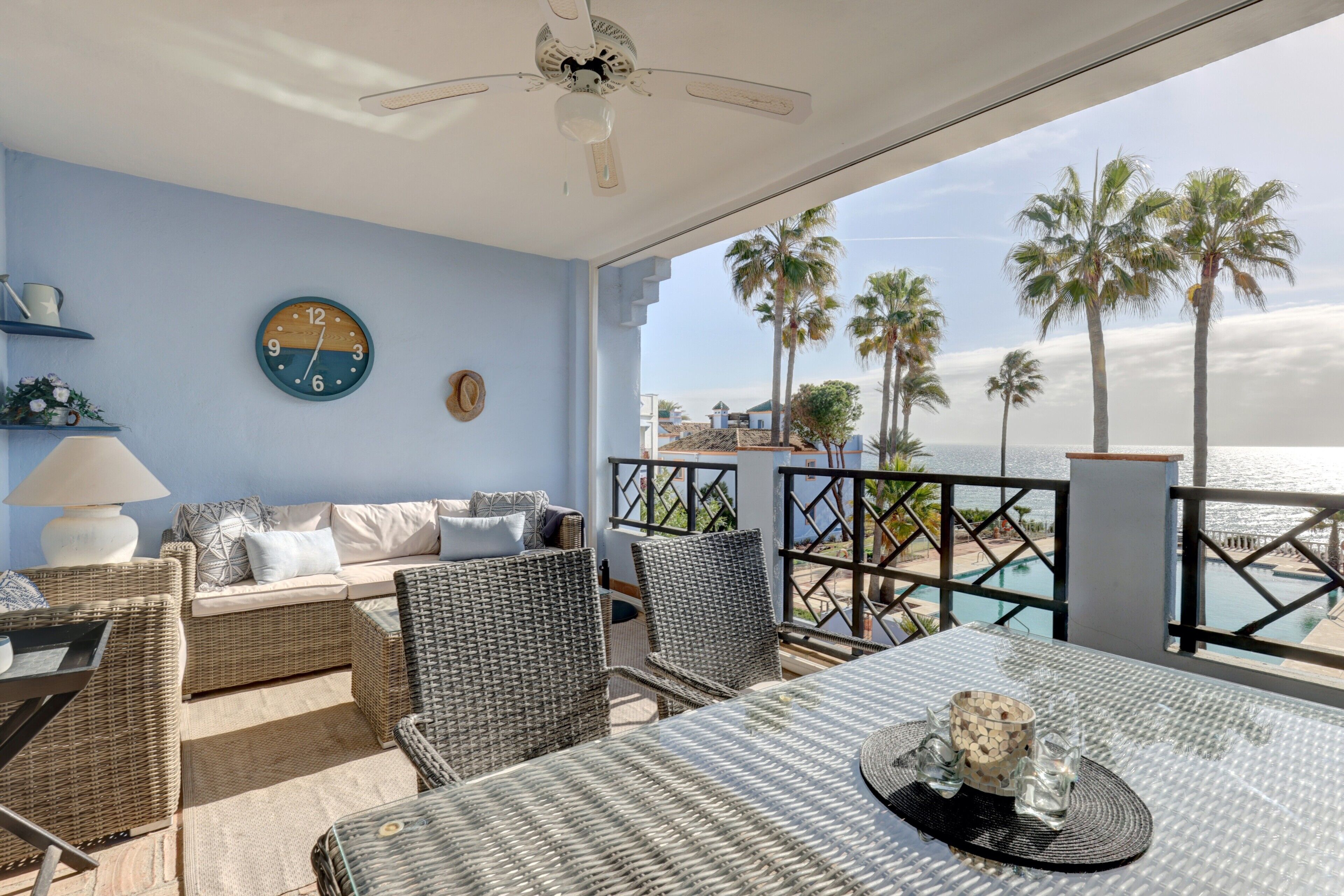 A balcony with wicker furniture, a clock, and a view of palm trees and a pool.
