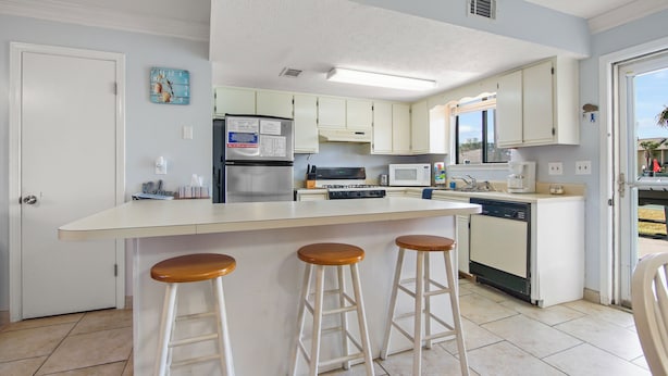 A modern kitchen with white cabinets, a stainless steel refrigerator, a microwave, a stove, and a dishwasher. There are three wooden stools at the island. - Your Beach Getaway Awaits YOU! (Panama City Beach)