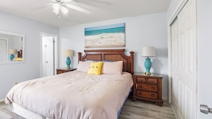A bedroom with a bed, two bedside tables, a ceiling fan, a painting of a beach, and a white door. - Your Beach Getaway Awaits YOU! (Panama City Beach)