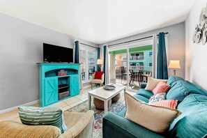Un salon doté d’un meuble de rangement pour appareils multimédias de couleur turquoise, d’un ensemble de canapés, d’une table basse et offrant une vue sur un balcon aménagé avec du mobilier d’extérieur.