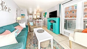 A living room with a teal sofa, white coffee table, and a kitchen area with a teal cabinet. - Harbor House 2BR 2BA Gulf-View 1st floor Wrap Balcony Pool King Bed (Gulf Shores)