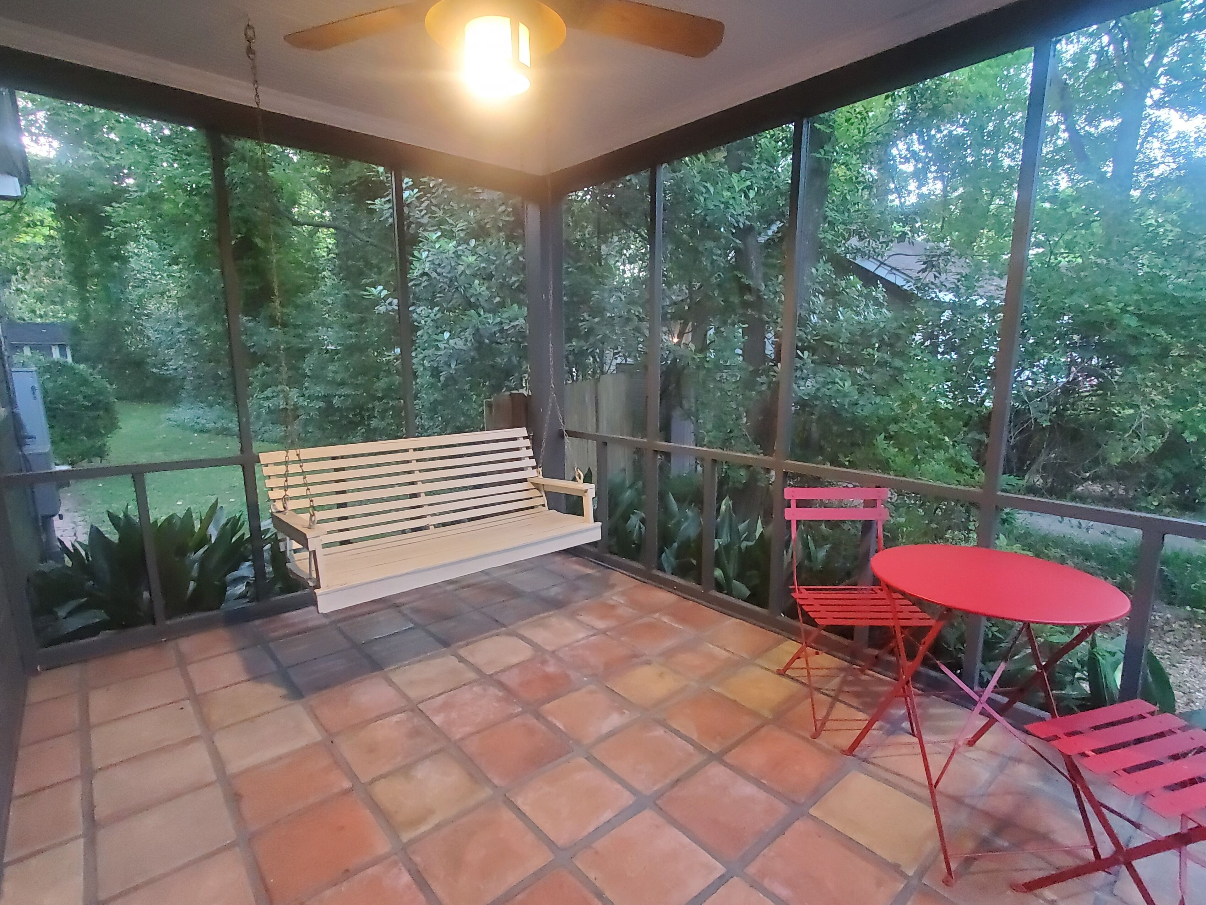Ein Balkon mit einer hölzernen Bank und roten Stühlen, einem gefliesten Boden und Blick auf einen Garten mit Bäumen.