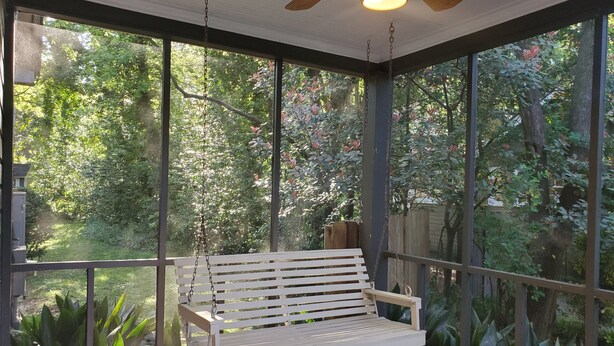 Eine überdachte Veranda mit einer weißen Schaukelbank, großen Fenstern und einem Deckenventilator.