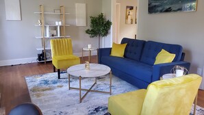 Ein Wohnzimmer mit einem blauen Sofa, gelben Sessel und einem runden Couchtisch sowie einem Bücherregal.
