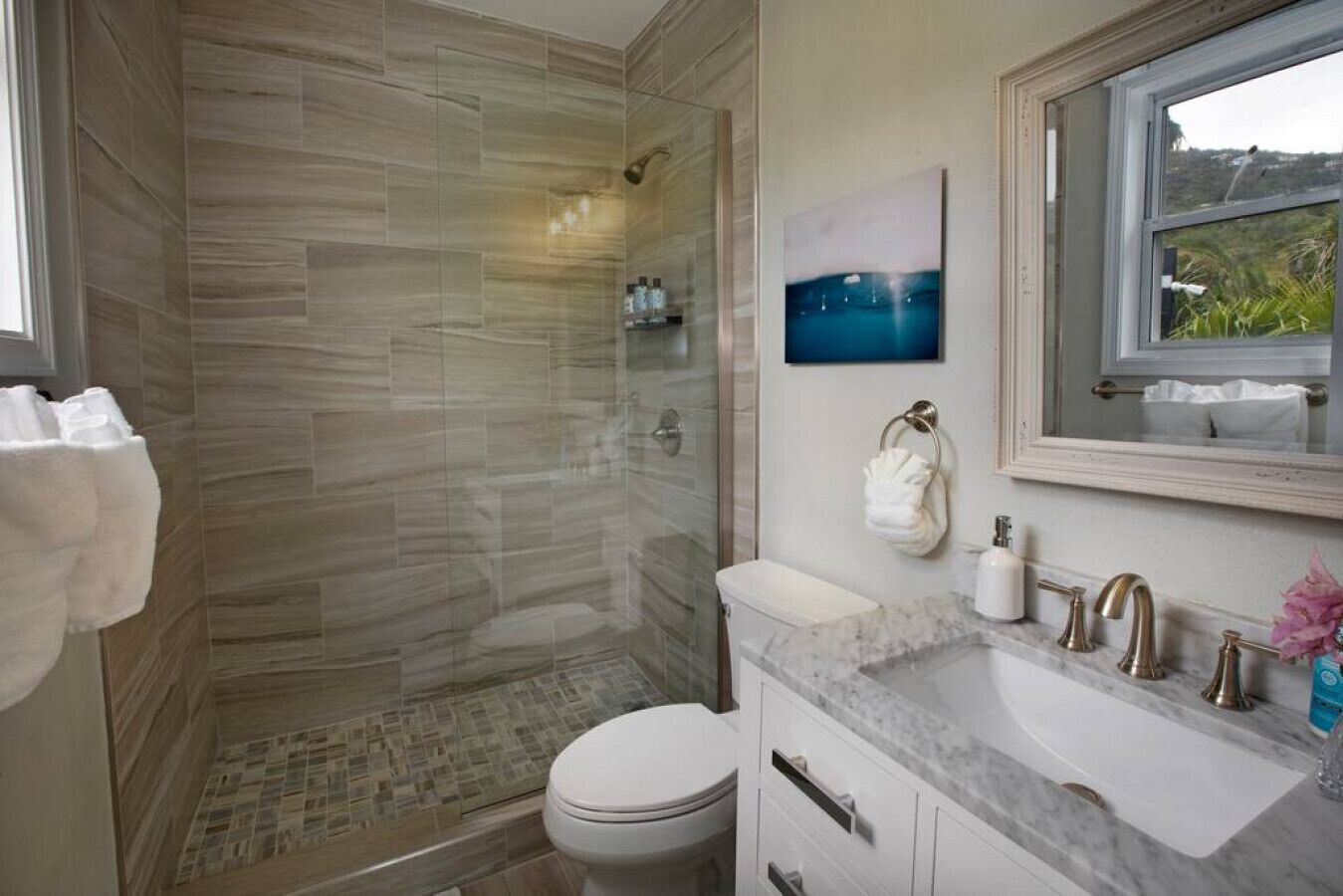 A modern bathroom with a glass shower enclosure, a toilet, a sink with a marble countertop, and a window with a view of greenery.