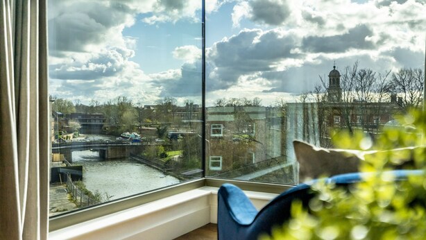 Et værelse med et stort vindue der kigger ud over en kanal og en bro, en stol og en potteplante.