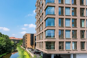 A modern multi-story building with a brick facade, large windows, and a river in the foreground. - Beautifully Furnished, Central, 2 Bedroom Apartment - With Parking (York)