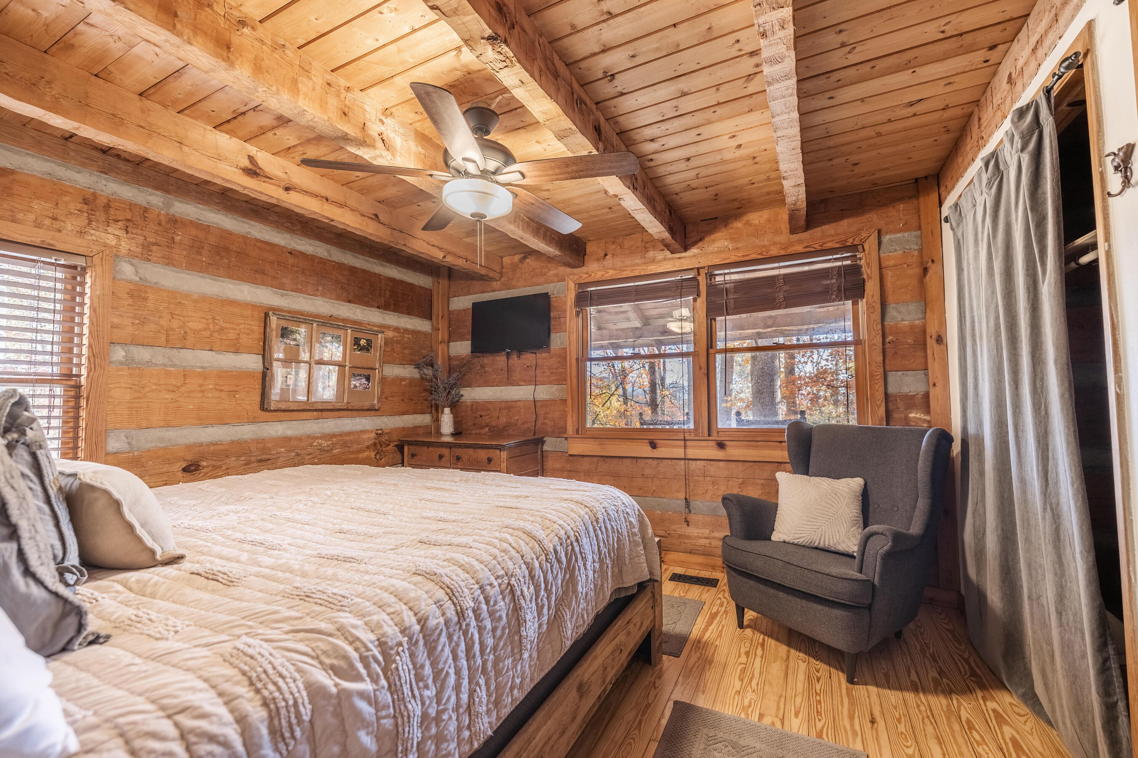 A cozy log cabin bedroom with a bed, a chair, a ceiling fan, and large windows.
