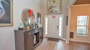 Um corredor com piso de madeira, um console de madeira escura com objetos decorativos, um espelho com uma fita vermelha e um quadro acima de uma porta com a inscrição 