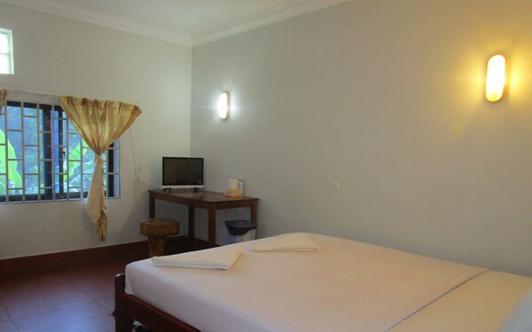 A hotel room with a bed, a desk with a television, a window with curtains, and a wall-mounted light.