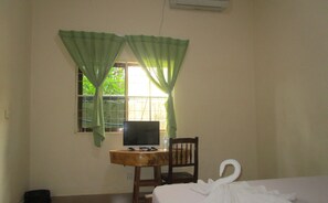 Una habitación con cama, escritorio con computadora, silla y ventana con cortinas verdes.