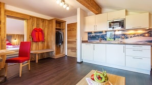 A modern room with a wooden floor, a red chair, a wooden bench, a dining table with a fruit bowl, and a kitchen with white cabinets and a microwave.