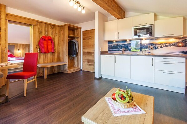 A modern room with a wooden floor, a red chair, a wooden bench, a dining table with a fruit bowl, and a kitchen with white cabinets and a microwave.