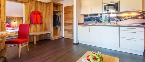 A modern room with a wooden floor, a red chair, a wooden bench, a dining table with a fruit bowl, and a kitchen with white cabinets and a microwave.