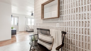 Una habitación con papel tapiz estampado, un espejo con marco de madera, un banco con cojín y vistas a un pasillo con una puerta y una planta.