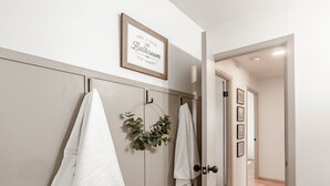 Un pasillo con una puerta que da a un baño, un cartel enmarcado sobre la puerta y una planta colgando de la pared.