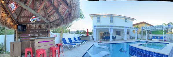 A pool area with lounge chairs, a bar, and a house with a patio. - 5 Mins to beach; 5BD, 4.5BA, Heated pool, Jacuzzi, Mini Golf, Tiki Huts.  (Boynton Beach)