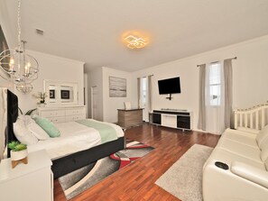 A modern bedroom with a bed, bedside tables, a desk, a TV, and a living area with a sofa and a rug. - 5 Mins to beach; 5BD, 4.5BA, Heated pool, Jacuzzi, Mini Golf, Tiki Huts.  (Boynton Beach)