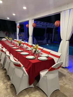 A long dining table set for a formal event with white chairs and red tablecloths, overlooking a pool area. - 5 Mins to beach; 5BD, 4.5BA, Heated pool, Jacuzzi, Mini Golf, Tiki Huts.  (Boynton Beach)