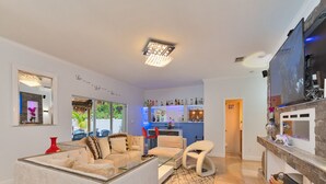 A modern living room with a sofa, armchairs, and a coffee table. There is a bar area with bottles and a TV mounted on the wall. - 5 Mins to beach; 5BD, 4.5BA, Heated pool, Jacuzzi, Mini Golf, Tiki Huts.  (Boynton Beach)