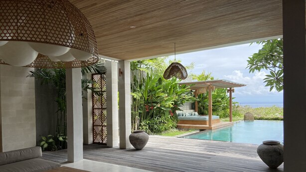 A modern outdoor lounge area with a wooden ceiling, a cushioned seating area, a wooden deck, and a view of a pool and ocean. - Villa Biarritz (Pecatu)