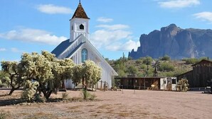 Une église blanche surmontée d’une croix, entourée d’une végétation désertique et avec une montagne en arrière-plan.
