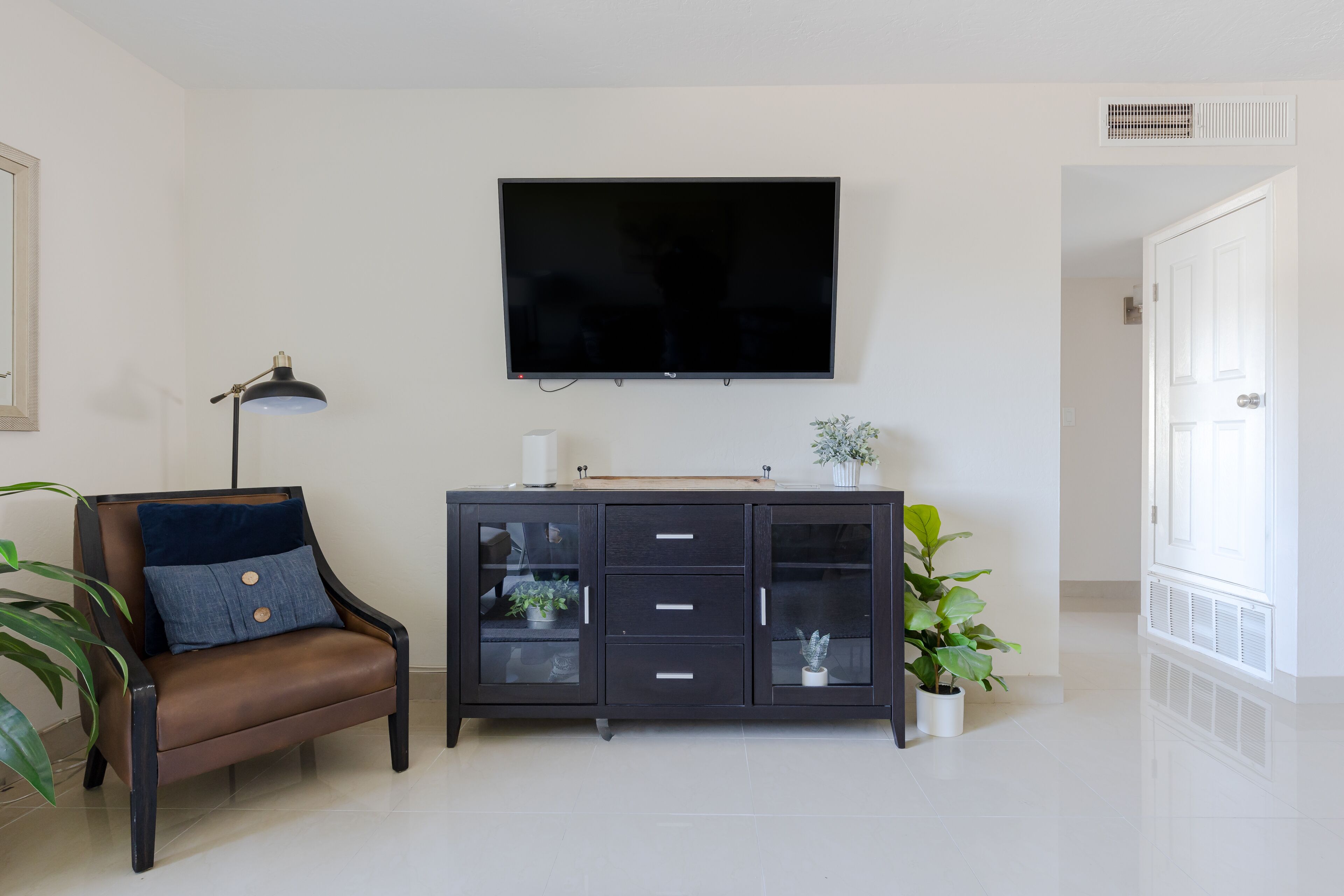 A living room with a flat-screen TV mounted on the wall, a dark wooden cabinet with glass doors, a brown leather sofa with a blue cushion, and a floor lamp.