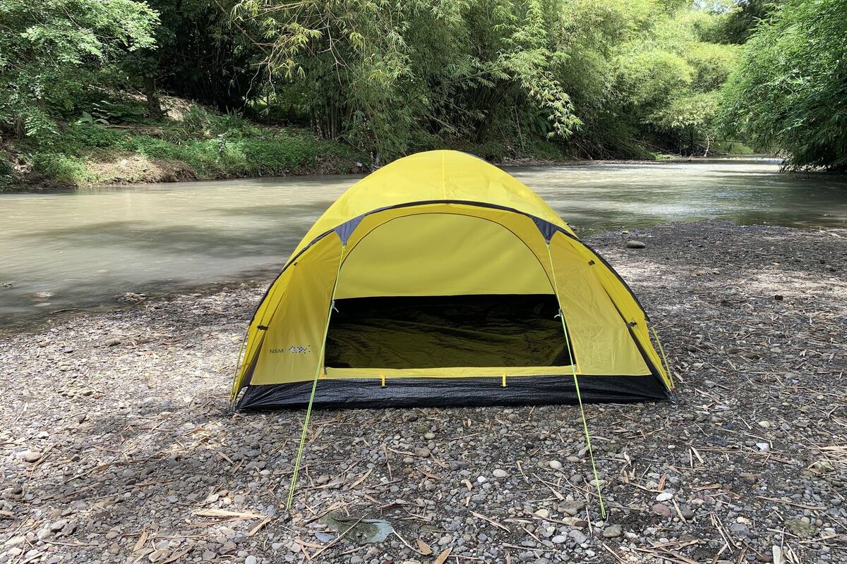 A yellow tent set up on a gravel path beside a river, surrounded by dense greenery.