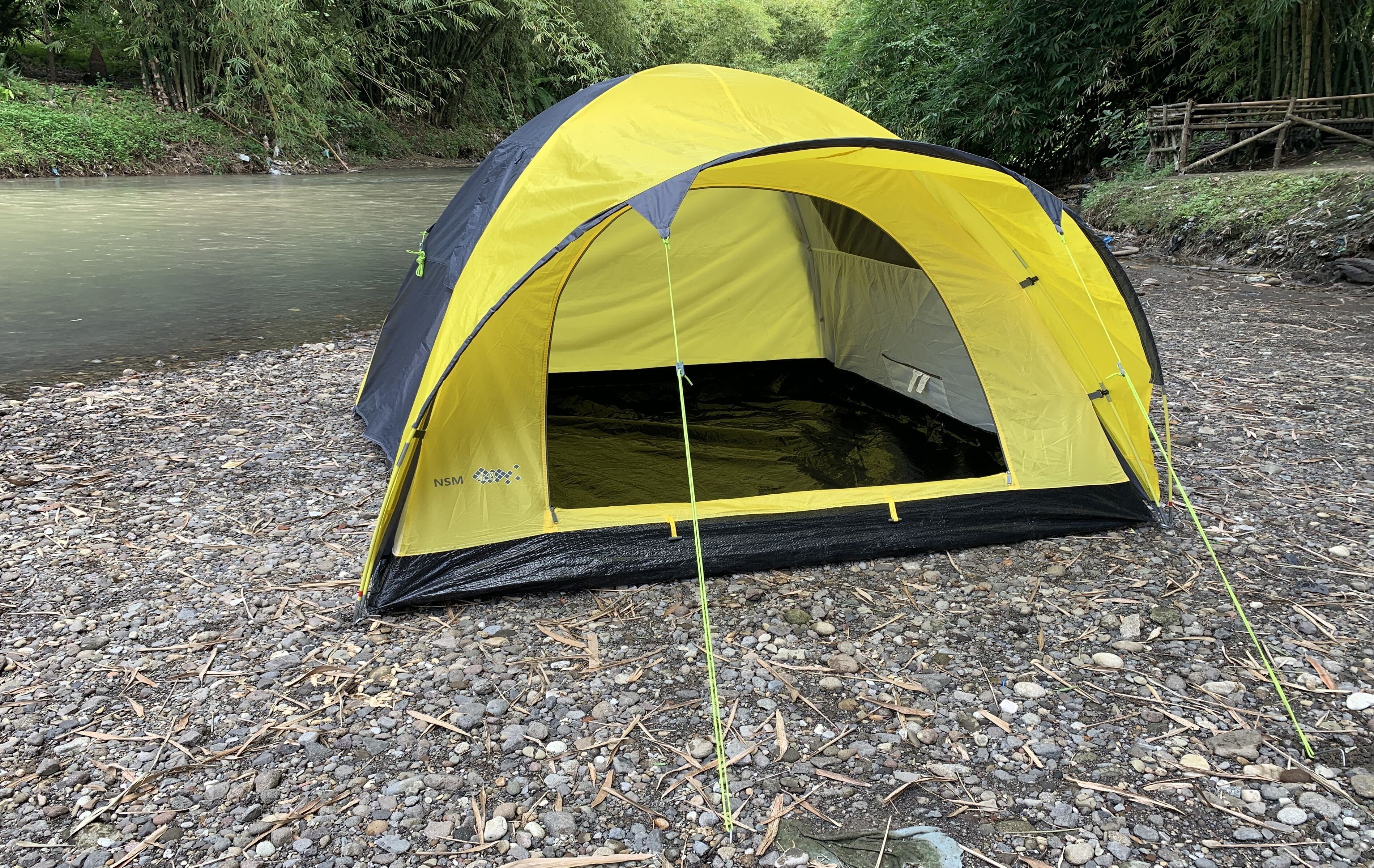 Une tente jaune et noire est installée sur une rive rocheuse, avec une rivière en arrière-plan.
