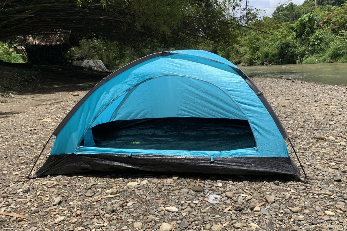 A blue and black tent set up on a gravel path near a river, surrounded by greenery.
