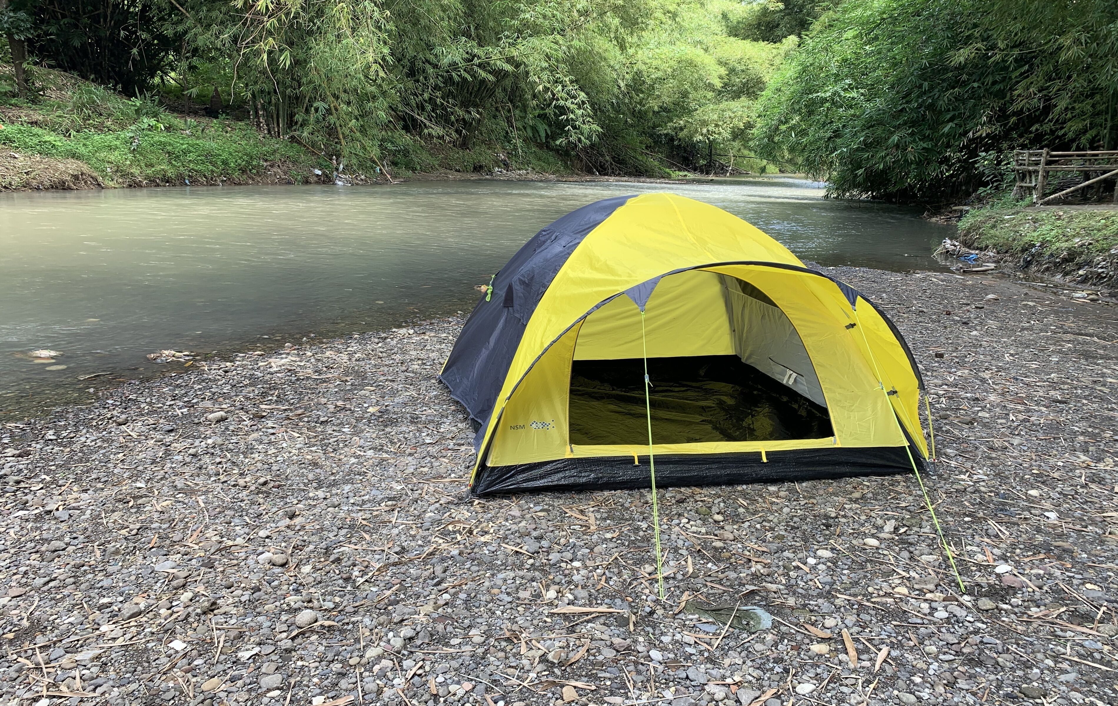 A yellow and gray tent set up on a gravel path beside a river, surrounded by dense greenery.