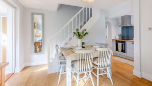 A dining area with a round table, white chairs, a chandelier, and a staircase leading to the kitchen. - Bright and cosy two bedroom holiday cottage with wood burning stove - Hillcrest Cottage (Avening, near Tetbury)
