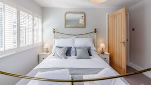 A bedroom with a bed, two bedside tables, a mirror, a wooden door, and a window with blinds. - Bright and cosy two bedroom holiday cottage with wood burning stove - Hillcrest Cottage (Avening, near Tetbury)
