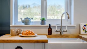 A kitchen with a window, a sink, a teapot, a plant, a bottle, and a croissant on the counter. - Bright and cosy two bedroom holiday cottage with wood burning stove - Hillcrest Cottage (Avening, near Tetbury)