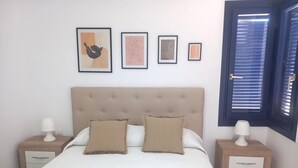 Un dormitorio con una cama grande, dos mesitas de noche, una ventana con persianas y tres cuadros en la pared.