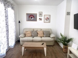 Een woonkamer met een beige bank, gestreepte kussens, een houten salontafel en een televisie aan de muur.