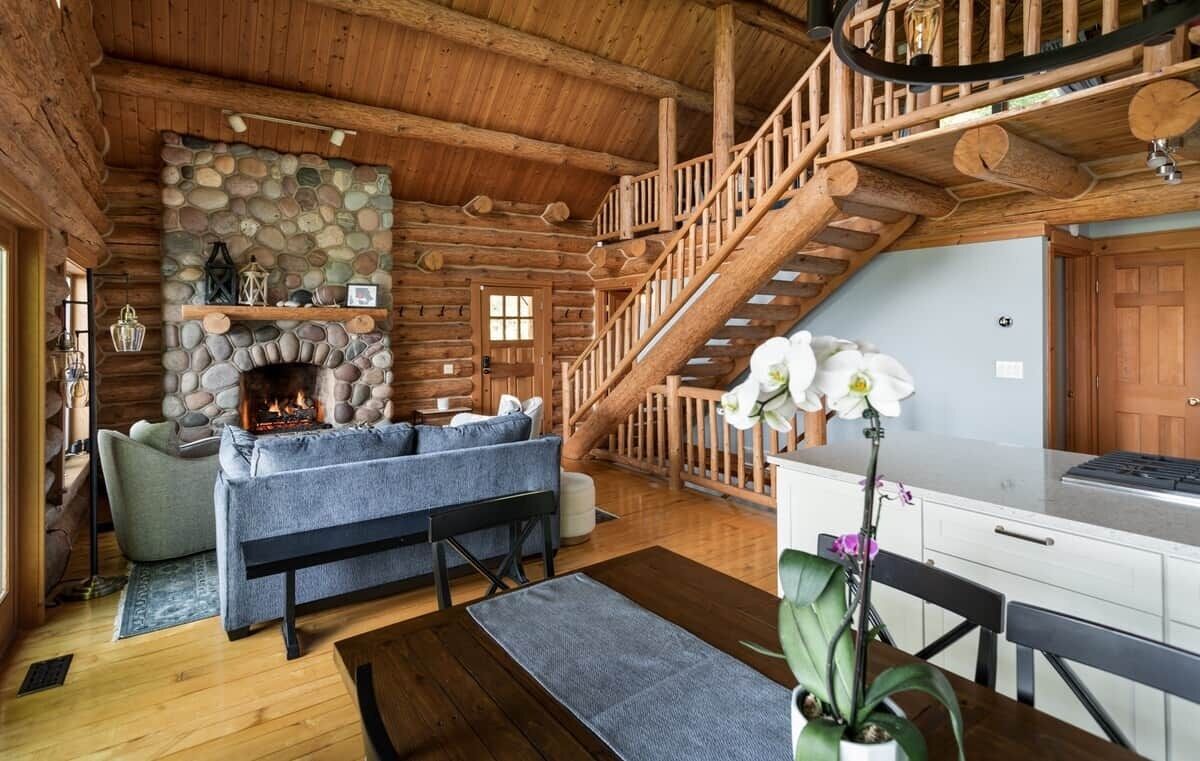 A log cabin interior with a stone fireplace, a staircase, a sofa, and a dining area.