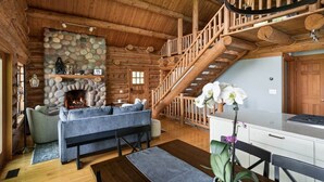 Interior de uma cabana de tronco com lareira de pedra, escada, sofá e área de jantar.