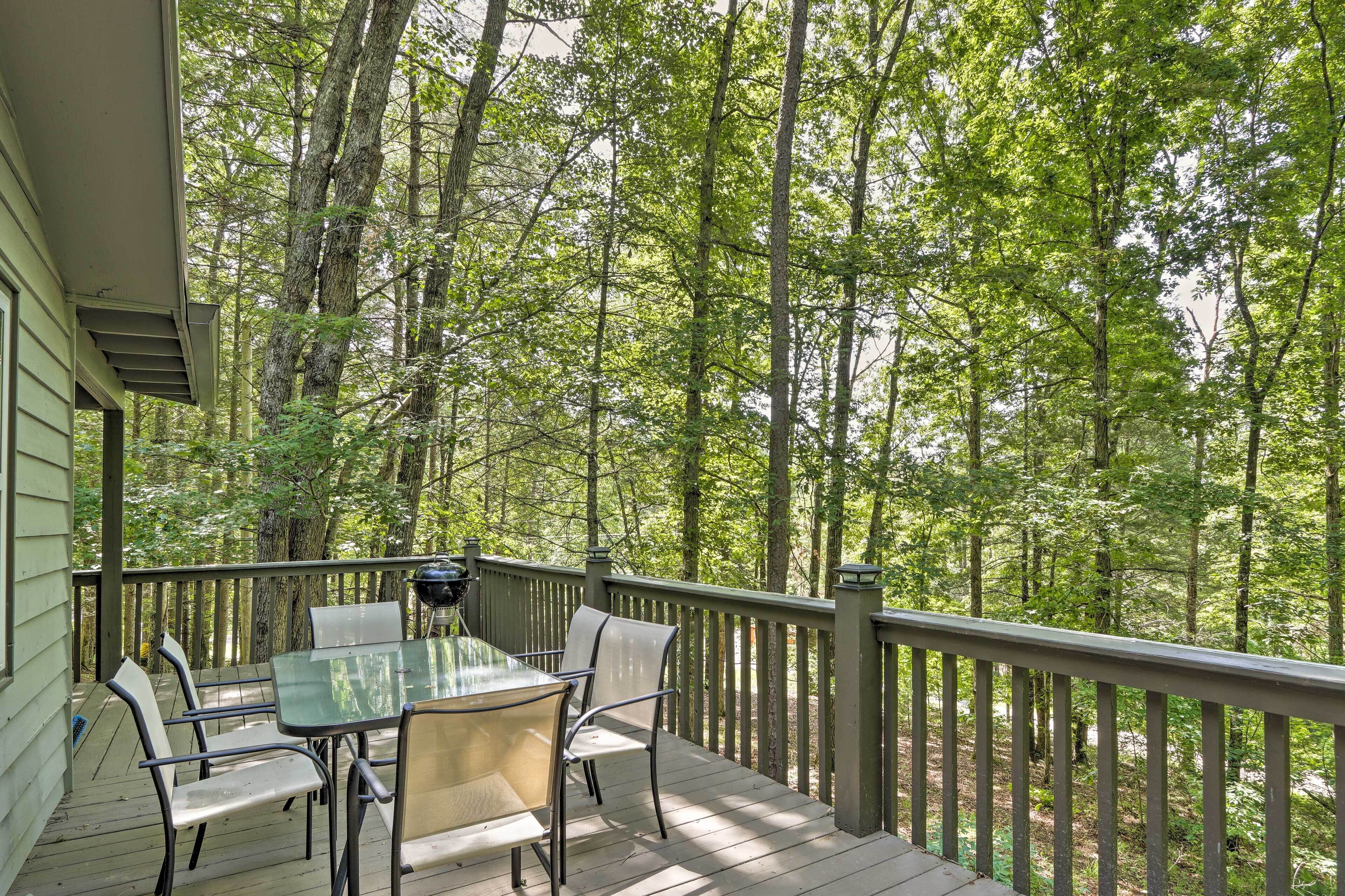 A wooden deck with a glass table and chairs, surrounded by a forest.