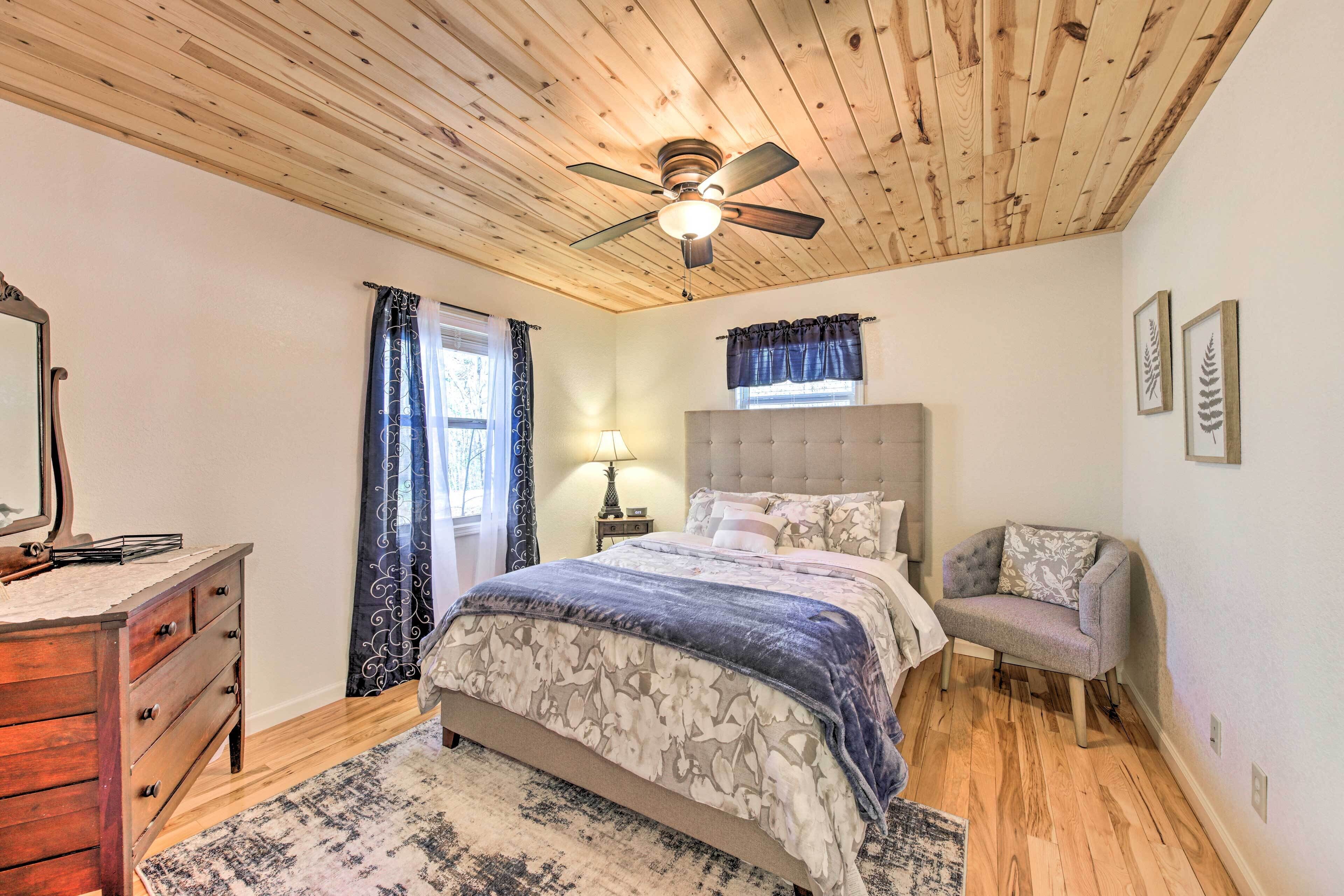 A bedroom with a wooden ceiling, a bed with a floral bedspread, a nightstand with a lamp, a chair, and a dresser.