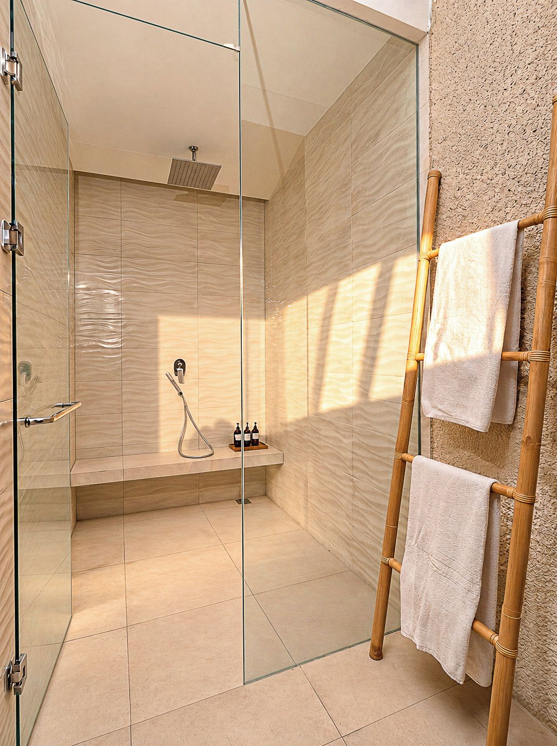 A modern bathroom with a glass shower enclosure, a wooden ladder with towels, and a bench with toiletries.