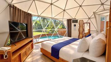 A bedroom with a bed, a TV, a wooden dresser, and a view of a pool and greenery.