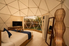 A tent-style room with a bed, TV, and a view of greenery outside. - Triyana Resort and Glamping (Carangsari)