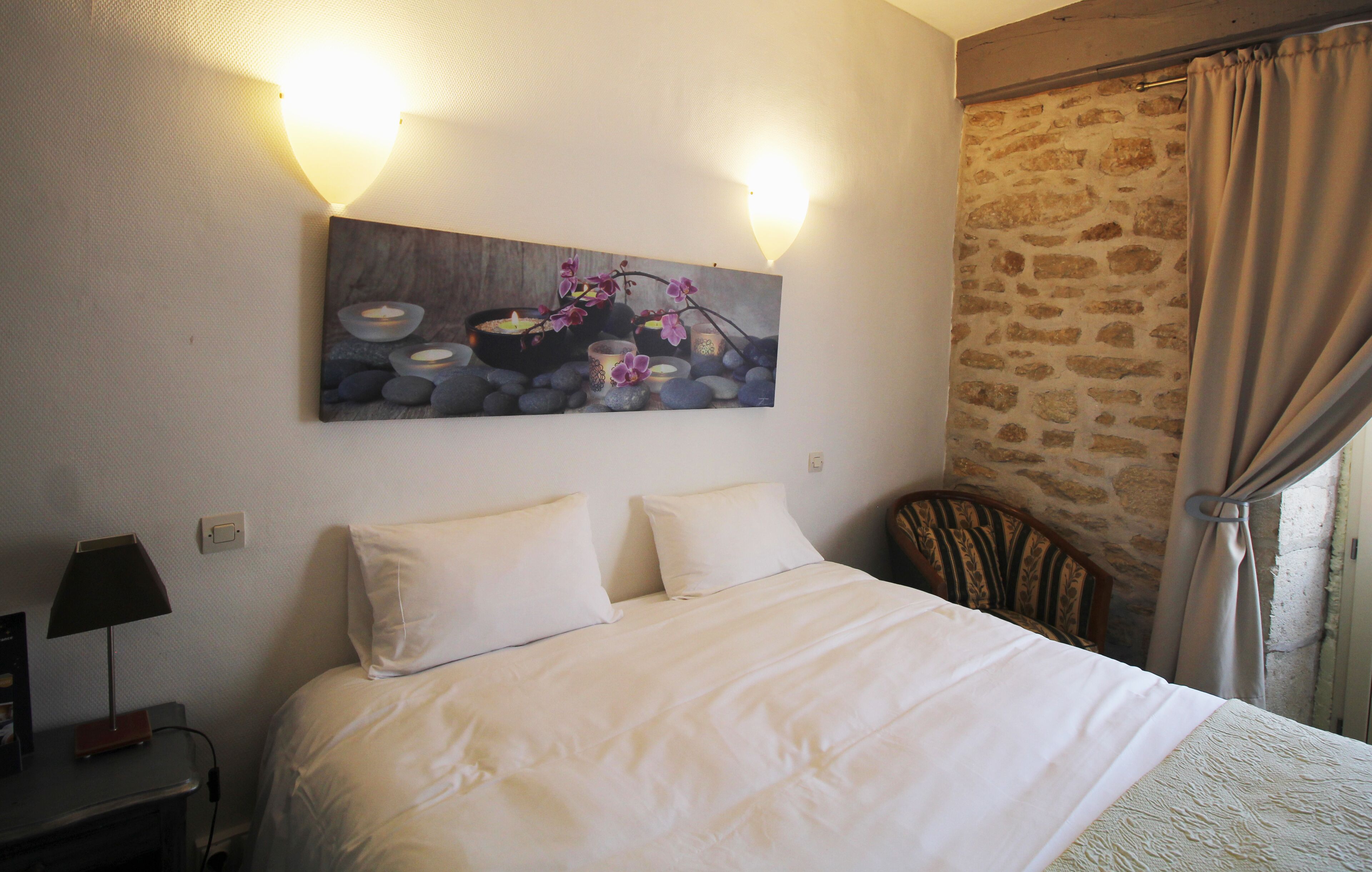Una habitación de hotel con una cama grande, dos lámparas de noche, una mesita de noche, un cuadro con flores y velas, y una pared de piedra.