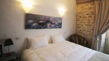 Una habitación de hotel con una cama grande, dos lámparas de noche, una mesita de noche, un cuadro con flores y velas, y una pared de piedra.