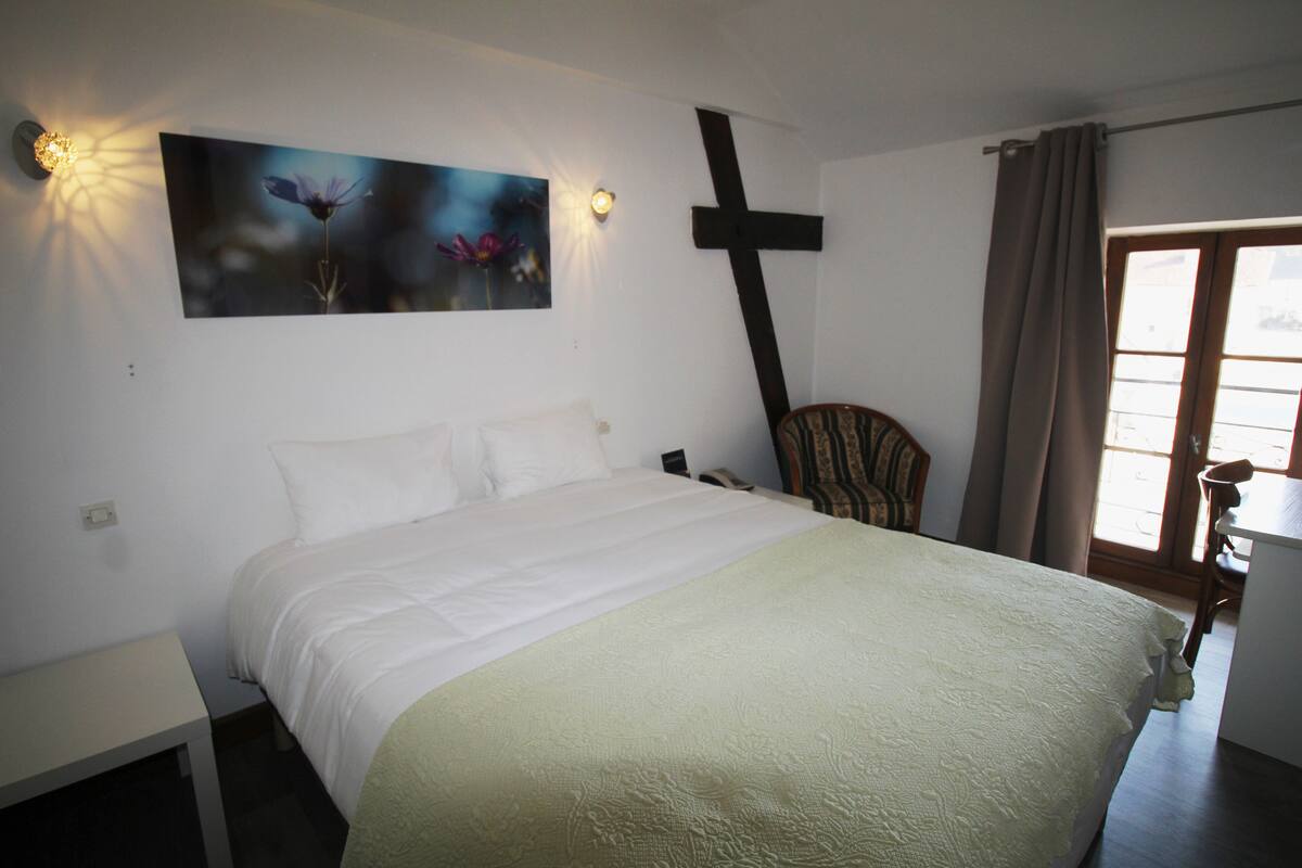 Quarto com cama, cruz na parede, cadeira e janela com cortinas.