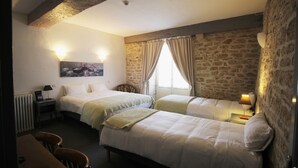 Ein Zimmer mit zwei Betten, einem Holztisch und einem Stuhl. An der Wand hängt ein Gemälde, auf dem Nachttisch steht eine Lampe und das Fenster ist mit Vorhängen drapiert.